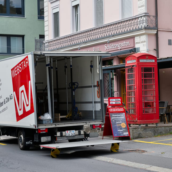A moving vehicle with a large open side panel is parked on a street in front of residential buildings, supporting a home relocation process. The vehicle is branded with 'WEBSTAR' and has a lowered hydraulic lift at the rear, with yellow and black stabilisers visible underneath for stability. Inside the vehicle, there are packing materials and tools used for furniture transport, including a blue hand truck and some white bags. To the right of the vehicle, a traditional red British telephone box is situated on the pavement, adjacent to an outdoor seating area with a small black table and chairs. The buildings behind feature pastel-colored facades, one with a decorative iron balcony, and a sign above indicates a pub called 'Schützengarten'. The scene captures the typical urban environment while highlighting the logistics involved in house removals and loadings performed by specialists like Man with Van Kensington.
