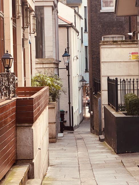 A narrow, paved alleyway between residential buildings in an urban area, with a small garden bed enclosed by a red brick wall on the left and a black metal fence on the right. Traditional black street