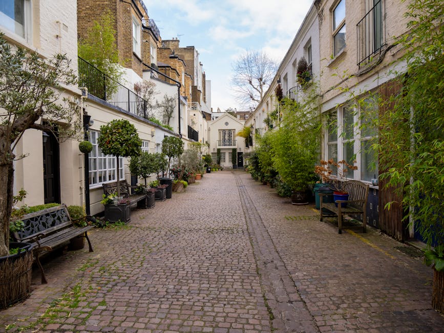 A narrow cobblestone street in Kensington, lined with red-brick Victorian-style buildings featuring small balconies with black iron railings. On the left, there is a shopfront with a green awning and