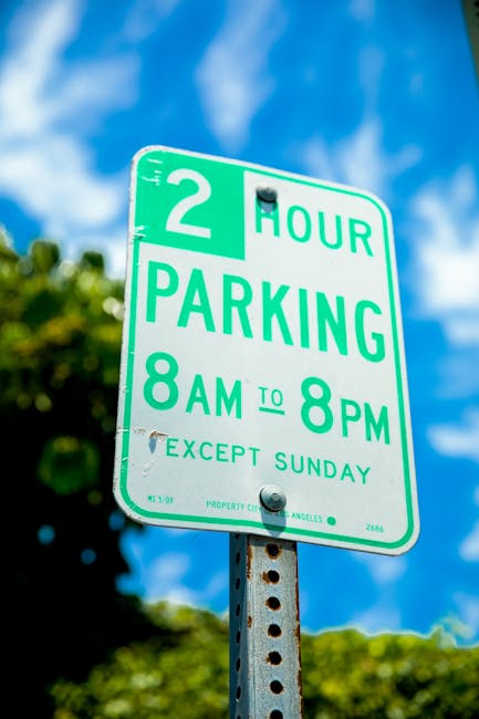 A close-up of a green and white parking regulation sign mounted on a metal pole against a background of a blue sky with some clouds and green leafy trees. The sign indicates a two-hour parking limit from 8 am to 8 pm, Monday to Saturday, with an exception for Sundays. Its placement suggests it is located in an urban residential area, likely within Kensington, where house removals and packing services by Man with Van Kensington might be performed. The sign's detailing, including the time restrictions and exception clause, reflects local council rules for parking permits relevant to moving logistics. The weather appears clear and sunny, providing good visibility for the sign, which is essential for drivers and residents managing home relocations or furniture transport in the area.
