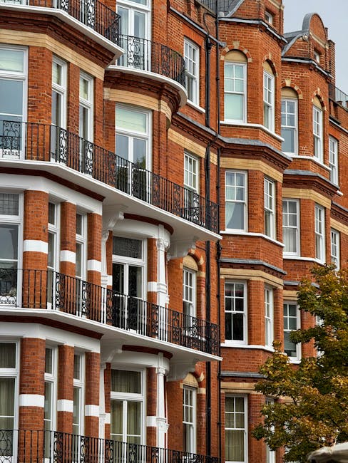 A multi-storey brick residential building with traditional Victorian-style architecture, featuring large white-framed sash windows and decorative white stone accents. The building has several curved and straight balconies with black wrought iron railings, some with glass doors leading to interior rooms. On the ground level, part of a pavement and a tree with green and yellow leaves are visible, indicating a street scene. The exterior is well-lit with natural daylight, highlighting the rich red brick and contrasting white detailing. This environment reflects an urban setting suitable for house removals and furniture transport, where typical logistics might involve packing items into cardboard boxes, wrapping furniture with blankets or plastic wrap, and loading them onto vans for home relocation. The building’s architecture is characteristic of Kensington in London, aligning with the services offered by Man with Van Kensington during moving and packing processes.