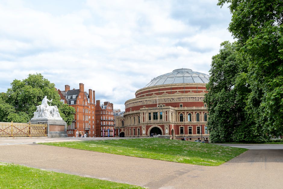 A wide view of the Royal Albert Hall in Kensington, London, with a clear sky overhead. In the foreground, there is a paved pathway and a section of green grass. To the left, a white stone monument featuring sculptures of seated figures is situated near a decorative wrought iron fence. On the right, large leafy trees frame the scene, partially obscuring the building. The photograph captures the iconic venue, often involved in various cultural and community events. This setting is relevant to home relocation and moving services in Kensington, as it highlights the surrounding environment and landmarks near the area where Man with Van Kensington provides house removals and furniture transport services.