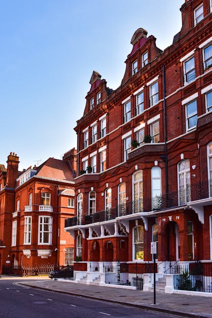 The image depicts a row of red-brick Victorian-style terraced buildings on a residential street in Kensington, with multiple large windows and ornate architectural details. In the foreground, a black van with its rear doors open is parked on the pavement, and the loading area is partially visible. Inside the van, several cardboard boxes, some wrapped with plastic film, are stacked, ready for a home relocation. A few boxes are being carried by a man dressed in casual clothing, who is loading or unloading them from the van. Near the open doors of the property, a trolley loaded with additional boxes and packing materials such as bubble wrap and moving blankets is visible. The scene is well lit by natural daylight, with a clear blue sky overhead, and the surroundings suggest an organized furniture transport and packing process typical of professional removals services, notably those offered by Man with Van Kensington, during a move in W8 Kensington High Street.