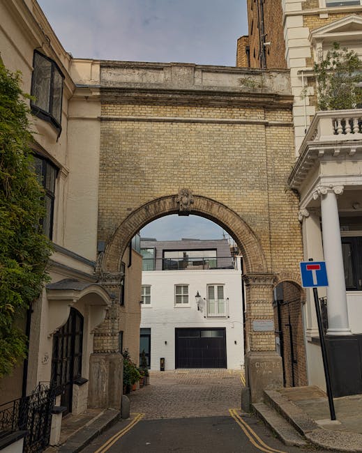 A narrow street in Kensington featuring a historic brick archway with a decorative crest at the top, leading into a courtyard or parking area. To the left, there is a cream-colored building with a rounded bay window and some greenery climbing the wall, while on the right, a white building with a classical facade, including columns and a balcony. A blue 'no through road' traffic sign is mounted on a pole near the sidewalk. The street surface is paved with small stones and has yellow line markings along the curb, indicating parking restrictions. The lighting suggests daytime with clear weather, and the overall scene reflects a typical urban residential environment suitable for home relocations and furniture transport logistics managed by companies like Man with Van Kensington, especially when dealing with parking access issues for removals in the area.