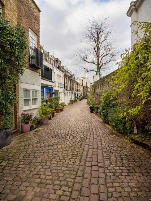 A narrow street in Kensington featuring a historic brick archway with a decorative crest at the top, leading into a courtyard or parking area. To the left, there is a cream-colored building with a rounded bay window and some greenery climbing the wall, while on the right, a white building with a classical facade, including columns and a balcony. A blue 'no through road' traffic sign is mounted on a pole near the sidewalk. The street surface is paved with small stones and has yellow line markings along the curb, indicating parking restrictions. The lighting suggests daytime with clear weather, and the overall scene reflects a typical urban residential environment suitable for home relocations and furniture transport logistics managed by companies like Man with Van Kensington, especially when dealing with parking access issues for removals in the area.