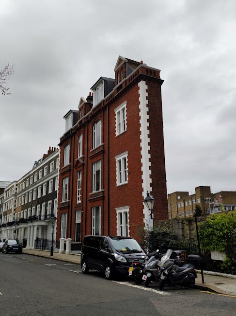 A multi-storey red brick building with white decorative accents on the corner of a street in Kensington, featuring several windows and a small rooftop structure under an overcast sky. The street in front shows parked vehicles, including a black van and several motorcycles, indicating a moving process or preparation for home relocation. Adjacent buildings are visible to the left, with some greenery behind the building, and a classic street lamp is mounted on the building's corner, supporting the urban residential environment. The scene reflects typical London architecture and conveys an area suitable for house removals and furniture transport services, as provided by Man with Van Kensington.