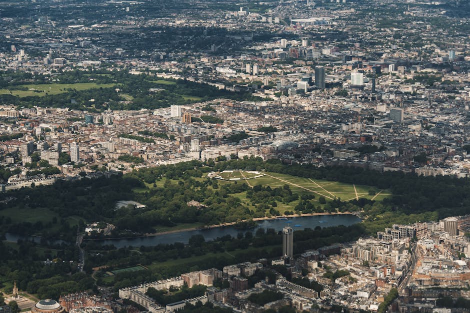 An aerial view of central London during daytime, showing vast green parks, including a large rectangular park with pathways and trees, bordered by dense urban buildings and streets. A river runs through the bottom part of the image, with a small bridge crossing it. The cityscape includes various high-rise buildings, residential areas, and commercial structures, with natural light highlighting the city's layout. This scene is relevant to home relocation and moving logistics, illustrating the urban environment where furniture transport or property transfers may occur, as seen from the perspective of a professional removals service like Man with Van Kensington highlighting the importance of planning in busy city areas.