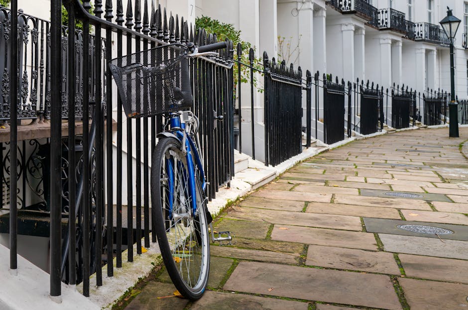 A blue bicycle with a black front basket is parked against a black wrought iron fence surrounding a white Georgian-style building with large windows and decorative columns on a residential street. The fence runs along a paved pavement made of large, irregular stone slabs with some grass and moss growing between the stones. Several black metal gates are evenly spaced along the fence, leading to the building's entrance steps. In the background, white balconies with black wrought iron railings and traditional lantern-style street lamps are visible. The scene is well-lit with natural daylight, and the overall setting suggests a quiet, upscale urban area suitable for home relocation or furniture transport services like those offered by Man with Van Kensington, with the fencing and steps indicating access points used during removals or packing and moving activities.