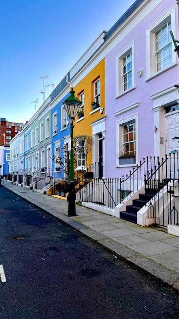 The image shows a row of brightly coloured terraced houses on a quiet residential street, with pastel facades in shades of purple, yellow, and blue. Each house has a small front garden enclosed by black metal railings and stone steps leading up to the front doors. Some houses feature window boxes and decorative elements above the windows, with clear glass panes and white frames. A traditional black lamppost stands on the pavement near the edge of the sidewalk, which is made of grey stone slabs. The street is paved with asphalt and appears clean, with parked cars visible further down the road. The scene is captured in daylight, with a clear blue sky and no traffic or pedestrians present, reflecting an orderly neighbourhood suitable for residential home relocation services. The image aligns with the theme of house removals and transportation, as it depicts typical urban architecture that a company such as Man with Van Kensington might service during furniture transport or moving logistics challenging areas like staircases and narrow doorways.